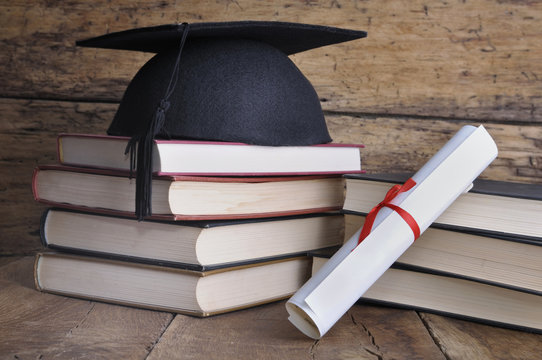 Graduate rolled up in a red ribbon and a black hat on pile of book on wooden  background