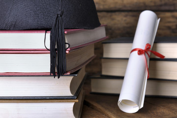 Graduate rolled up in a red ribbon and a black hat on pile of books on woden  background