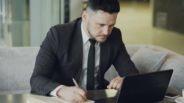 Concentrated bearded Caucasian businessman sitting and writing down business information in his notepad while looking at laptop in mod cafe