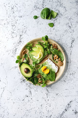 Vegetarian sandwiches with avocado, ricotta, egg yolk, spinach, walnuts on whole grain toast bread on ceramic plate with ingredients above over white marble background. Top view, space