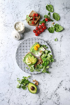 Vegetarian sandwiches with avocado, ricotta, egg yolk, spinach, cherry tomatoes on whole grain toast bread on ceramic plate with ingredients above over white marble background. Top view, space