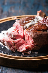 Sliced grilled sous-vide black angus beef tomahawk steak on bone served with salt, pepper, rosemary and white sauce on round wooden slate cutting board over dark wooden plank background. Close up