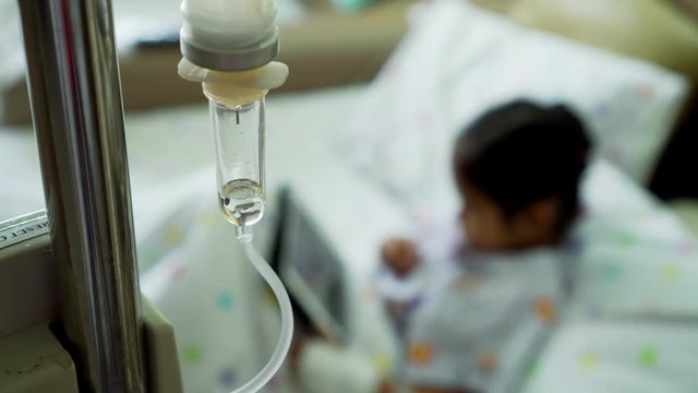Close up saline solution drip for patient with sick little child using digital tablet in the hospital blurred background. Patient in the hospital is receiving a saline solution by dripping machine.