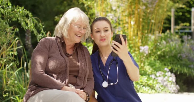 4K Young nurse taking care of senior lady, both posing for selfie in the garden on a sunny day. Slow motion