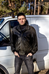 A young guy of criminal appearance in a black leather jacket stands near an old white van