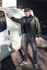 A young guy of criminal appearance in a black leather jacket stands near an old white van