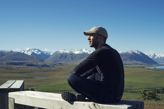 Hombre joven sentado en la cima de una montaña observando paisaje montañoso en Nueva Zelanda, 