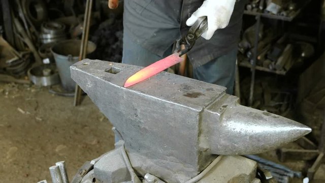 Making the knife out of metal at the forge. Close up blacksmith's hands hitting hot metal with a massive hammer on an anvil.