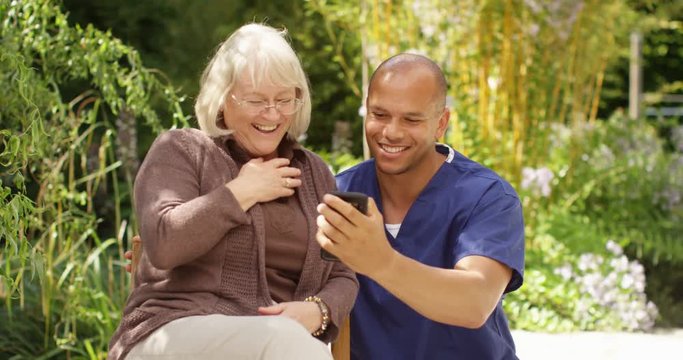 4K Happy mature lady laughing with her caregiver, looking at photos on a smartphone. Slow motion