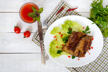 Fried pork ribs on a plate with greens and tomato juice on a white wooden background.