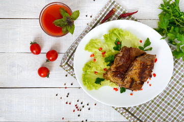Fried pork ribs on a plate with greens and tomato juice on a white wooden background.