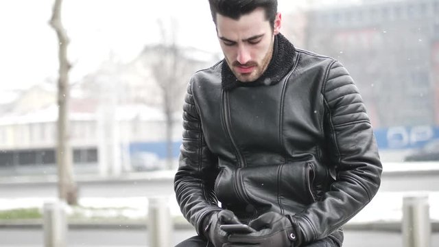 Handsome trendy man wearing black leather jacket sitting and typing on cell phone, outdoor in city setting in winter day shot