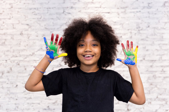 African American playful and creative kid getting hands dirty with many colors - in white brick background.
