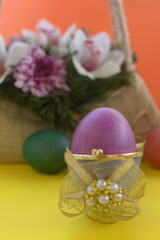 Purple egg in box with golden ribbon and pearl in front of linen basket with purple and white flowers and green and orange eggs in front on orange and yellow background 