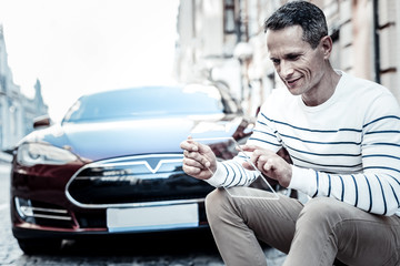 Technological device. Joyful nice smart man sitting near his car and smiling while using his tablet