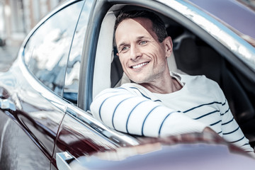 Great mood. Happy delighted positive man sitting in his car and smiling while looking into the window