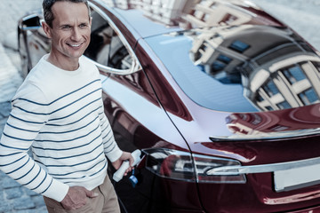 Great mood. Cheerful nice good looking man standing near his car and smiling while holding a petrol nozzle