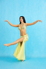 Charming dancer performs oriental belly dance on a blue background. A girl in a yellow beautiful dress is dancing an oriental belly dance on a blue background.