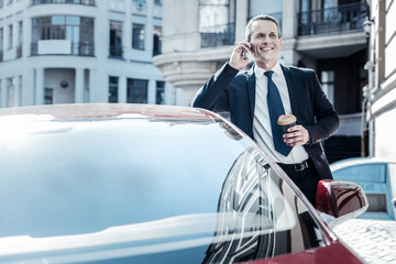 Favourite drink. Handsome pleasant nice businessman standing near his car and holding coffee while talking on the phone