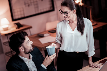 Have a rest. Pleasant bespectacled pretty employee standing in the office giving a cup of tea her colleague and smiling.
