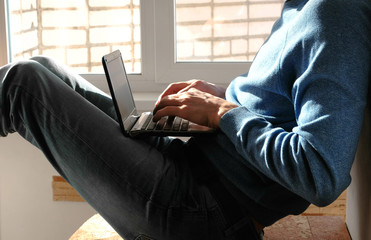 Working on laptop. Unrecognizable young man typing something in his laptop sitting on balcony near the window.