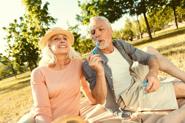 Fun activity. Nice pleasant aged man blowing soap bubbles and having fun while being on a picnic with his wife