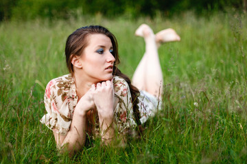 Naklejka na ściany i meble A beautiful brunette in a dress and a long scythe with a desired facial expression, lying on her stomach in the grass relaxes. The girl enjoys nature.