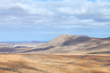 Fototapeta na wymiar Aerial view of arid desert valley with volcano peaks, coast village, on a summer, cloudy day, Fuerteventura, Canary Islands, Spain .