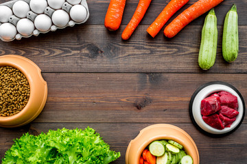 Dry pet food with natural ingredients. Raw meat, vegetables zucchini and carrot near eggs on dark wooden backgroud top view copy space