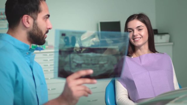 Good-looking doctor demonstrating x-ray to beautiful caucasian patient, young man wearing blue scrubs and holding teeth photograph in right hand, happy woman looking attentively
