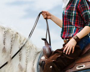 The cowboy on a white horse in western style holds the reins on a blue sky background isolated. Detail closeup