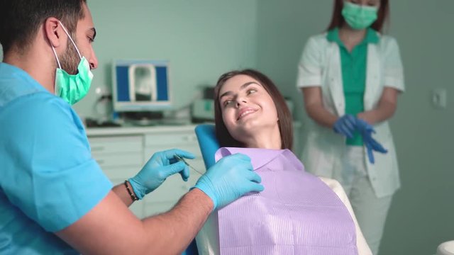 Cute young dentist putting purple bib on the patient, tall and slim assistant putting on rubber gloves, joyful patient talking to the doctor, indoor shot in large dental office