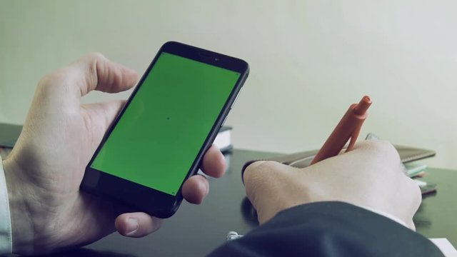Hands of businessman working in office at  with mobile green screen. One hand holds mobile phone with other hand, making notes with pen in notebook. Close-up of businessman hands with mobile phone.