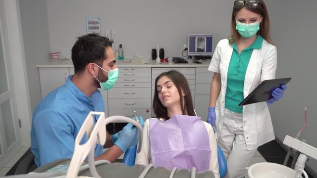 Dentist's assistant, in white uniform, brings black tablet, cute boy in blue uniform talking to attractive girl with a purple bib on the chest, doctors showing the patient treatment related images