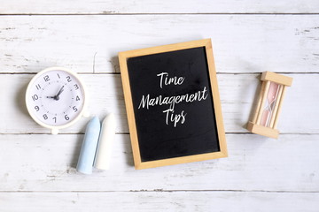 Top view of chalk,table clock,hourglass and blackboard written with 'TIME MANAGEMENT TIPS' on white wooden background.
