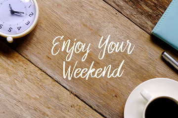 Top view of a cup of coffee,clock,notebook and pen on wooden background written with ENJOY YOUR WEEKEND.