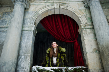 Brunette girl in green fur coat against old arch with columns and red curtains.