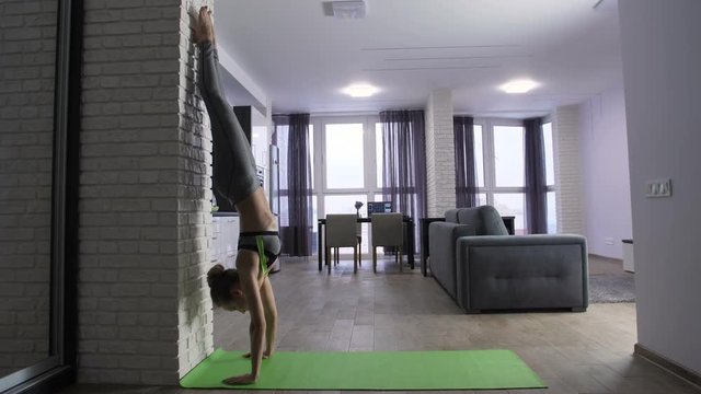 Full length of young fit attractive woman doing handstand near the wall at home. Morning exercise and gymnastics routine of slim mid adult female in sportswear. Healthy girl working out