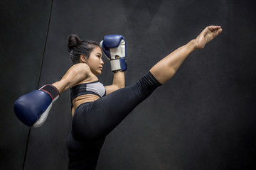 Jonge Aziatische vrouwenbokser met blauwe bokshandschoenen die in de oefeningsgymnastiek schoppen, vechtsporten op zwarte achtergrond