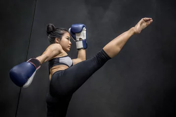 Photo sur Plexiglas Arts martiaux Jeune boxeuse asiatique avec des gants de boxe bleus donnant des coups de pied dans la salle d& 39 exercice, arts martiaux sur fond noir