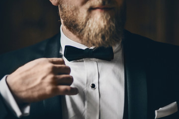 trendy bearded man in a black tuxedo adjusts his tie in the neck