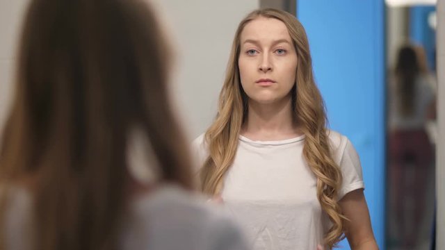 Depressed young woman looking at her portrait reflection in mirror. Beautiful mid adult woman with long blond hair displeased with her appearance. Body negative female sad and anxious about her look