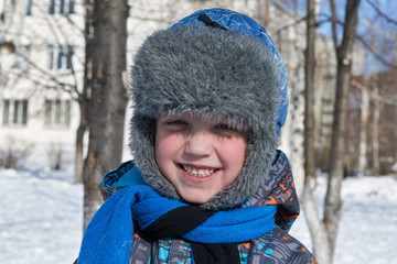 A small a boy winter sunlight a day to looks at us and smiles. On the street white snow. A boy a satisfied, joyous and happy. The child is dressed in a winter hat with fur, jacket and blue scarf.