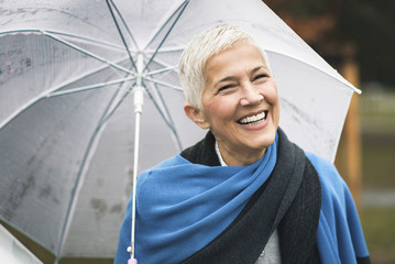Happy senior woman enjoying rainy weather outdoors in a park, holding an umbrella - 195554863