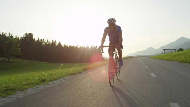 SLOW MOTION, SUN FLARE: Golden evening sun rays illuminate young athletic man pedaling his cool road bicycle. Caucasian cyclist enjoying picturesque ride on his bike through the sunny countryside.
