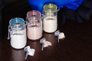Three glass jars with protein powder, strawberries, banana and chocolate and three spoons for milkshakes on the background of sortive accessories, Boxing gloves, bandages for brushes and shaker