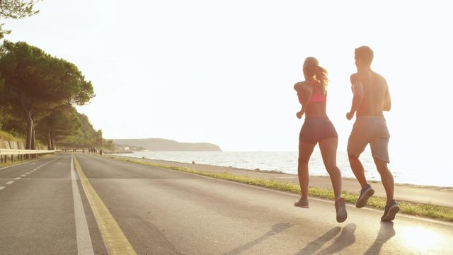 SLOW MOTION, LENS FLARE, COPY SPACE: Unrecognizable jogging couple on holiday soaking up the sun rays. Healthy couple running down beautiful sunny road overlooking the ocean. Relaxing evening jog.