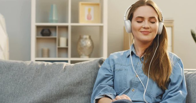 Portrait shot of the smiled young woman in the jeans shirt and white headphones enjoying of listening to the music on her smartphone with eyes closed while sitting on the couch in the cozy apartment