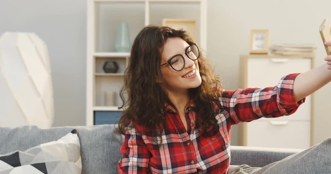 Portrait of of the young curly woman in the glasses and plaid shirt making a selfie on the smartphone on the sofa in the cozy room. Indoor