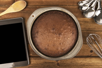 Freshly baked chocolate cake, tablet and utensil disposed on wooden table. Top view.
